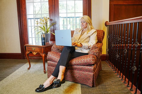 Woman in business attire sitting in an upholstered armchair using a laptop in an elegant room with windows and wooden furniture.
