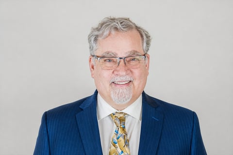 Professional headshot of a middle-aged man with gray hair and beard, wearing glasses, navy pinstriped jacket, and patterned tie against a light background