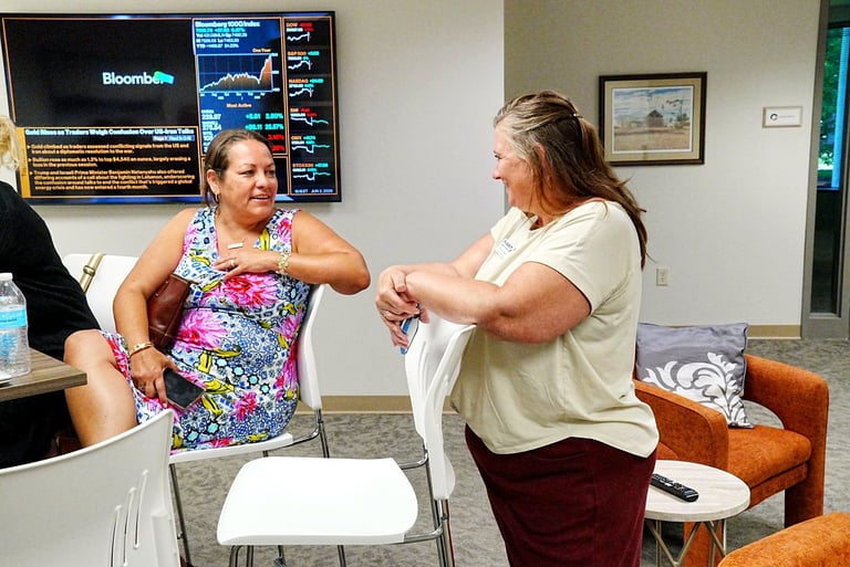 Two women greet each other with an elbow bump in an office setting with Bloomberg display and furniture visible in background