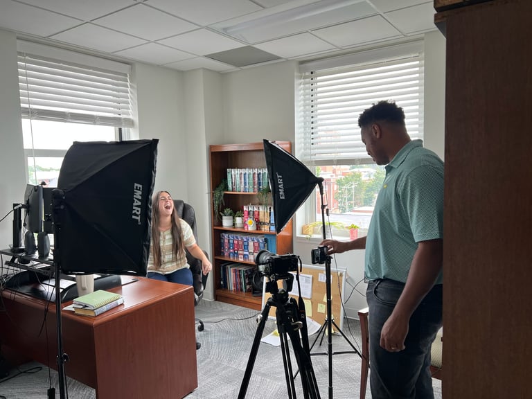 Two people conducting a video or photo shoot in an office setting with professional lighting equipment and camera on tripod.