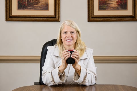 Professional woman with blonde hair sitting at desk in office, wearing white blazer and holding binoculars, with framed artwork on wall behind her