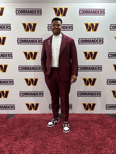 Man in burgundy suit and white dress shirt standing on red carpet at Washington Commanders media event