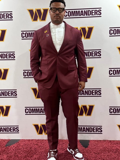 Man in burgundy suit and glasses at Commanders event backdrop with red carpet