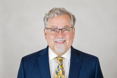 Professional headshot of a middle-aged man with gray hair and glasses wearing a dark pinstriped suit and patterned tie