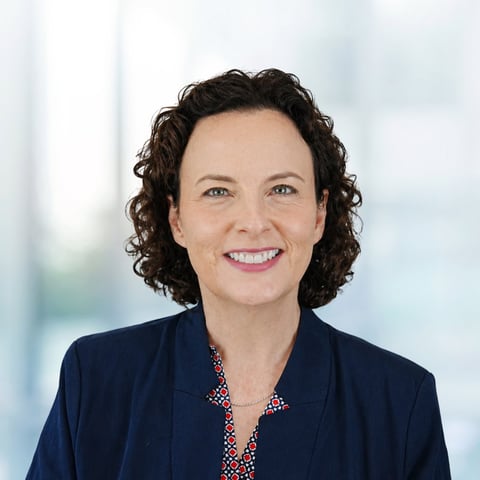 Professional portrait of a woman with dark curly hair wearing a navy blazer and patterned blouse, smiling against a blurred office background