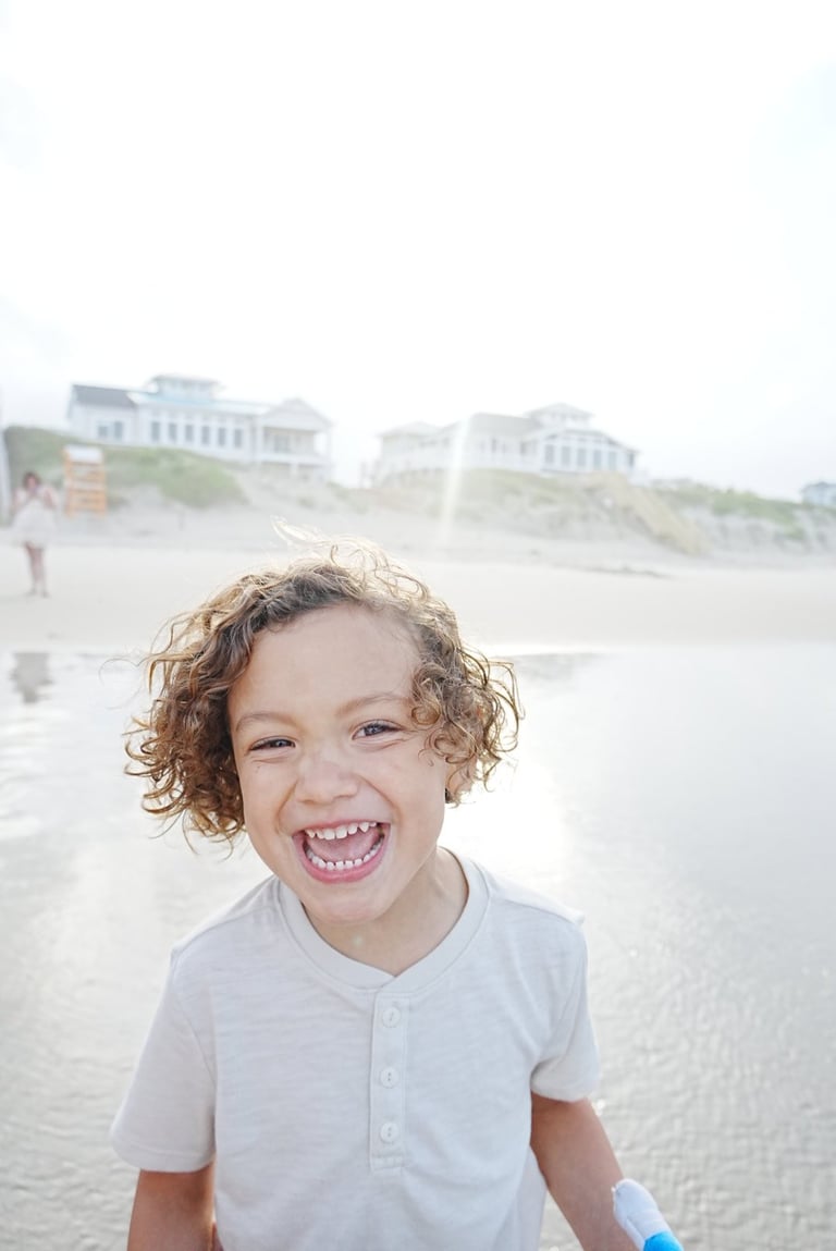Young child with curly hair smiling at the beach with beachfront houses in the background