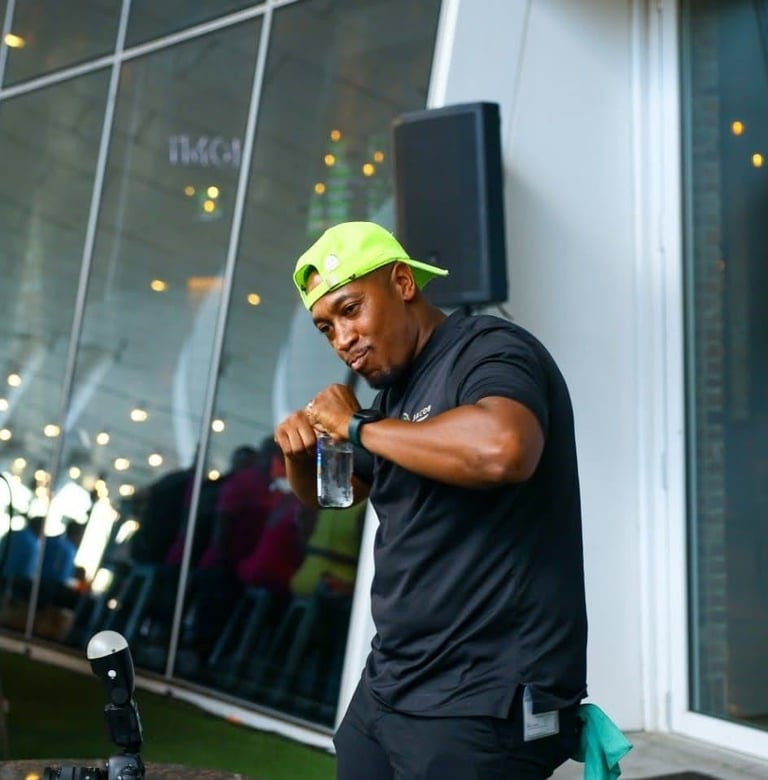 Man in neon yellow cap and black shirt holding a water glass, standing against glass building windows