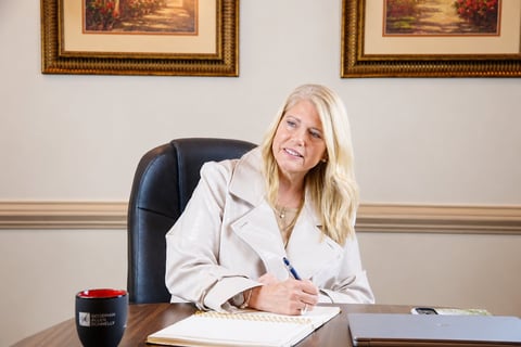 Professional woman with blonde hair in white blazer seated at desk with notepad and coffee mug in office setting