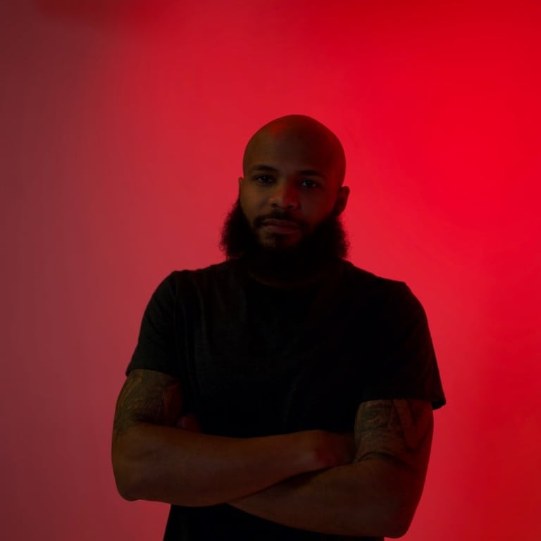 Portrait of a man with beard and tattoos on his arms, wearing a black t-shirt against a red gradient background