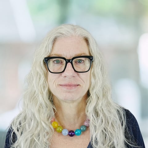 Portrait of a person with long white hair, black glasses, and a colorful beaded necklace against a blurred background