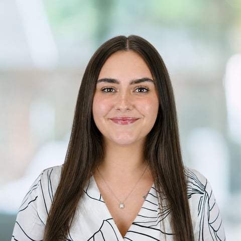Professional headshot of a young woman with long dark hair wearing a striped blouse, smiling at the camera with a blurred office background