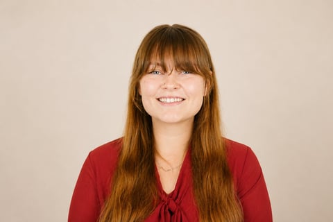 Woman with long reddish-brown hair and bangs wearing a red top, smiling at the camera against a neutral beige background