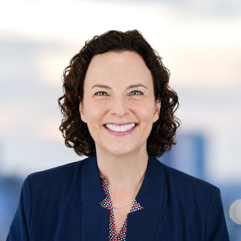 Professional headshot of a woman with dark curly hair wearing a dark blazer and colorful blouse, smiling at camera with blurred office background