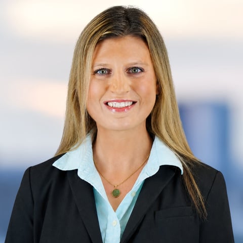 Professional headshot of a woman in business attire with long brown hair, smiling at the camera with a blurred cityscape background