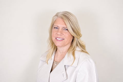 Professional headshot of a woman with blonde wavy hair wearing a white lab coat, smiling at the camera against a neutral background