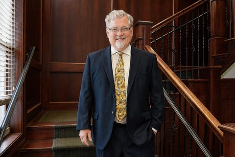 Older man with gray hair and glasses wearing a navy blazer and patterned yellow tie standing on a wooden staircase landing