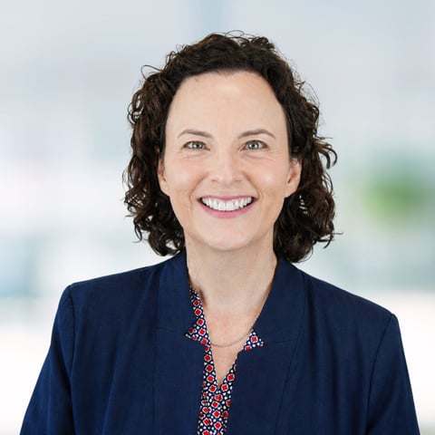 Professional headshot of a woman with dark curly hair wearing a navy blazer and colorful patterned blouse, smiling at the camera