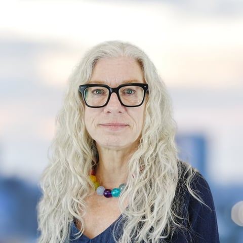 Woman with long white hair and black-framed glasses wearing a colorful beaded necklace against a blurred urban background