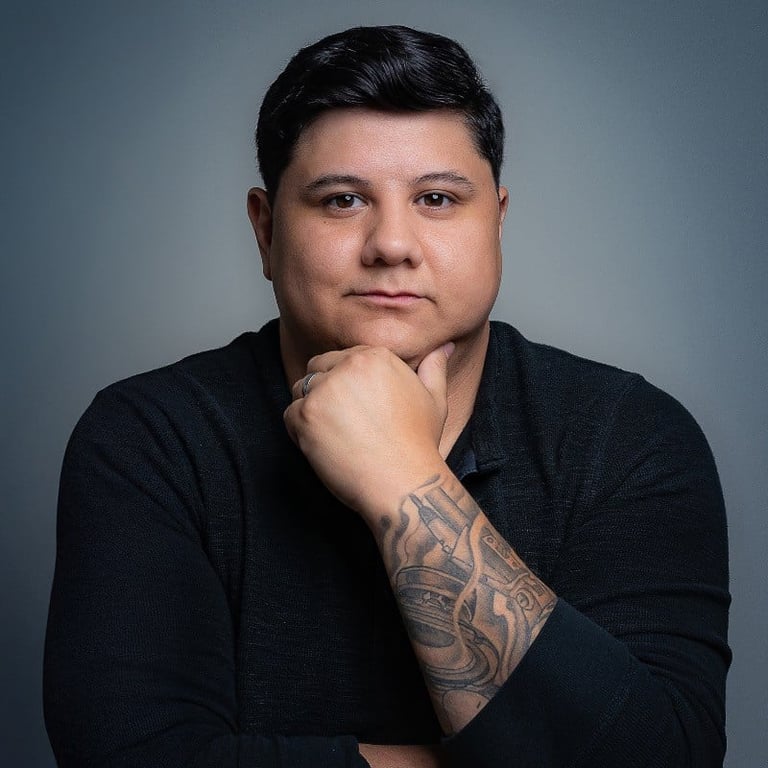 Man with dark hair and tattoo on arm posing with hand to face against gray background