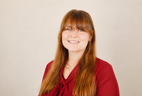 Portrait of a smiling woman with long reddish-brown hair and bangs wearing a red top against a neutral background