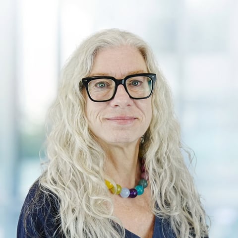 Woman with long white hair and black-framed glasses wearing a colorful beaded necklace, smiling at camera