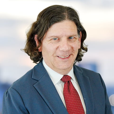 Professional headshot of a man with shoulder-length dark hair wearing a blue suit jacket and red tie, smiling at camera