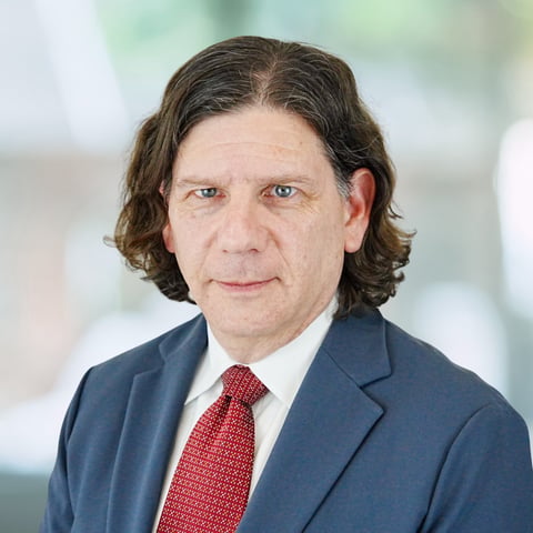 Professional headshot of a man with shoulder-length dark hair wearing a navy blazer and burgundy tie