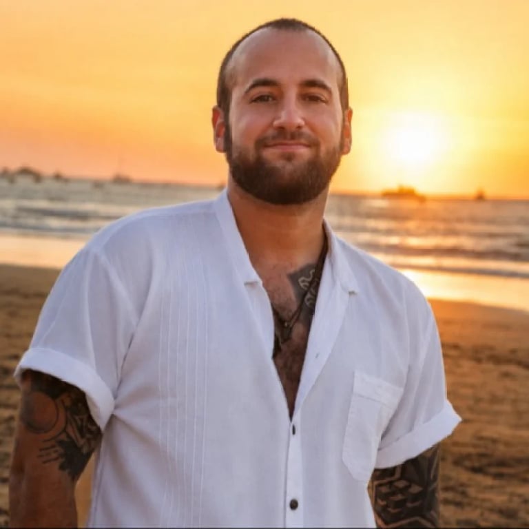 Man with beard wearing white short-sleeve shirt standing on beach at sunset