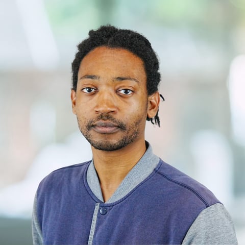 Professional headshot of a man with short dreadlocks wearing a navy and gray baseball jacket against a blurred light background