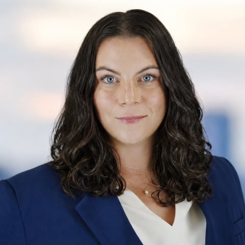 Professional headshot of a woman with long dark wavy hair wearing a navy blazer and white top against a blurred urban background