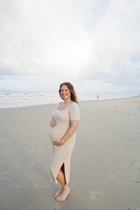 Pregnant woman in beige dress posing on beach with ocean waves and cloudy sky in background