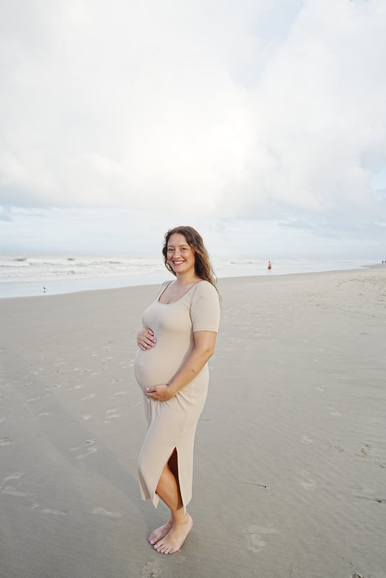 Pregnant woman in beige dress posing on beach with ocean waves and cloudy sky in background