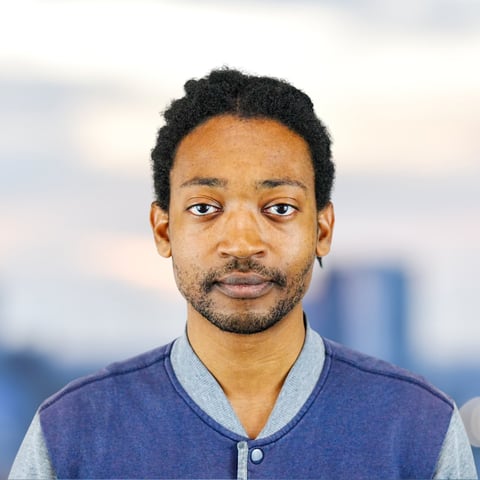 Portrait of a man with dark hair and facial hair wearing a blue and gray baseball-style jacket against a blurred city background
