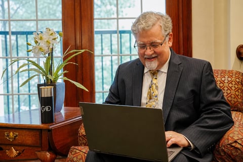 Older man in business attire with glasses working on laptop at wooden desk with flowers, formal interior with windows