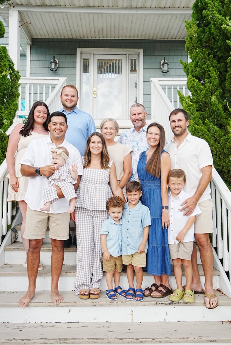 Multi-generational family of nine posing together on the front porch of a white beach house with gray siding