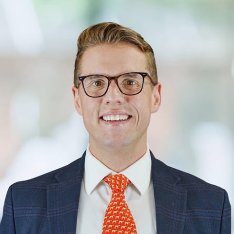 Professional headshot of a smiling man wearing glasses, navy suit, and orange tie