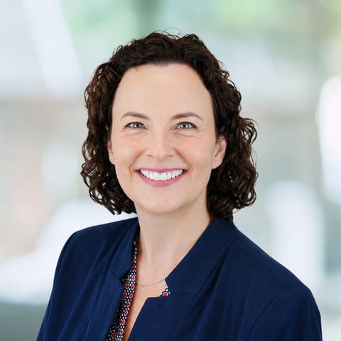 Professional headshot of a woman with dark curly shoulder-length hair wearing a navy blazer and patterned blouse, smiling at the camera